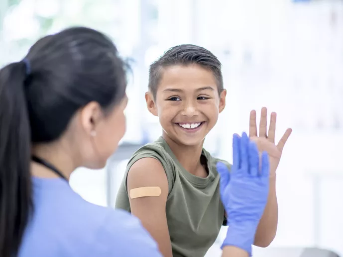 A young mixed-race boy visits a female doctor. The boy has a bandage on his arm where he received a vaccine injection. He is about to high-five the female doctor.